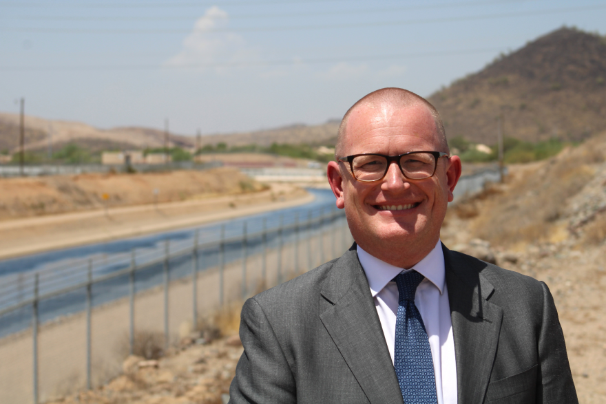 A man in a suit smiles at the camera near a flowing canal in an arid environment.