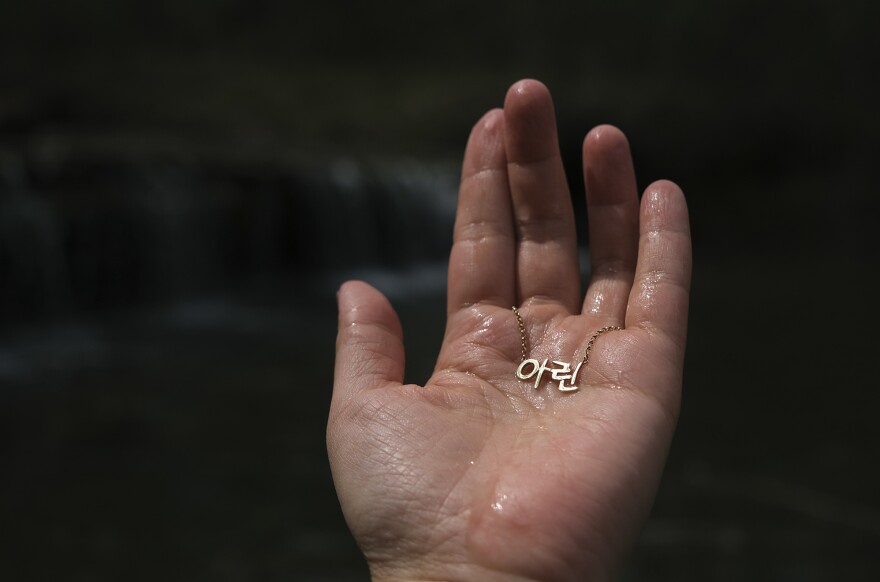 In her conception dream, Young Ok Na, the photographer's mother, holds up jewels from the stream to the light. She thinks of the Korean character 아<em> </em> (pronounced 'Ah'), which stands for beauty, and ë¦° (pronounced 'Lhin'), which is the sparkle of a jade. In an interpretation of that moment, Arin Yoon holds up her Korean name necklace to the sunlight.