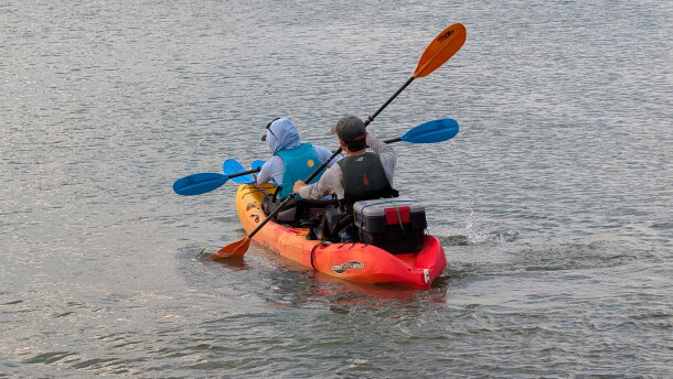 two people sit in a two-person orange kayak paddling out to sea. They have a box attached to the back of the kayak