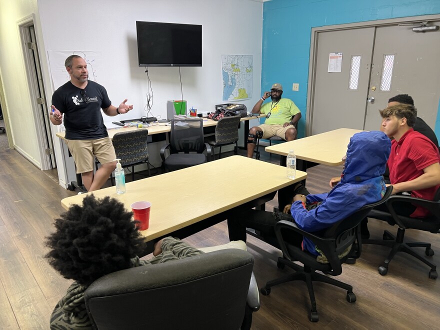 A group of teens sit at tables inside a community center. Two adults are in the front of the room speaking to them.