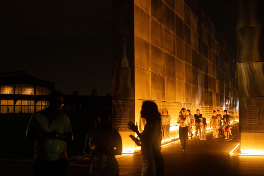 People walk and stand near a warmly lit memorial wall at night. Silhouettes and reflections are visible, with a dark building in the background.