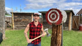 A lumberjack stands next to a throwing axe target while holding a double sided axe.
