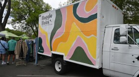 A classic box truck that’s painted with colorful swirls. On its left are two clothing racks with clothes and people walking alongside it. 