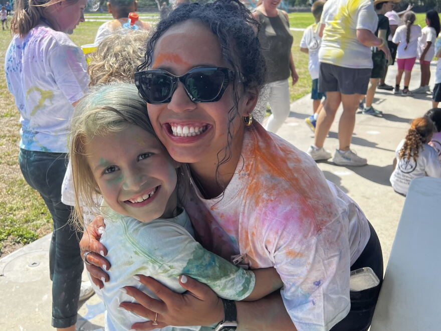 A woman and child, both covered in colorful dust, hug each other smiling at the camera. They are standing in front of multiple people outside on grass.