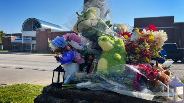 Outdoor photo shows a pile of flowers and stuff animals collecting on top of a concrete pillar. Behind, across the street, is a school building.