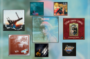 Artwork for four albums and three singles displayed in black square frames over a blurry blue-green background. In the top row from left to right: A person carrying a marimba on their back with the sun setting behind them, a hazy blue-green tinted photo of a woman wearing a red bandana, two people holding an upright bass and acoustic guitar inside a gold frame with red around it. In the bottom row from left to right: A woman in a dress with her arms above her head, a woman in a dark room with sun coming through a window, a pot on a hot plate with steam rising from it, and hard to make out red and white image of an amp with foliage around it.