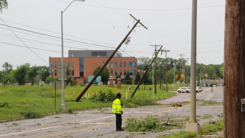 A man surveys the damage on a closed road. Leaves and branches are scattered around. Two power poles are at 45 degree angles. 