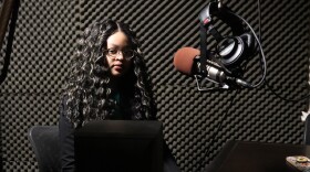 A woman with long curly hair and glasses sits in a soundproof radio studio, facing a microphone.