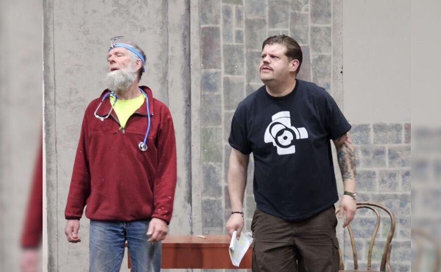 Two guys standing in front of a table and a grey brick wall.