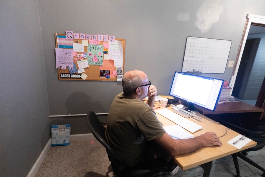 Austin Chronicle staff writer Brant Bingamon sits at a desk and looks at a computer. On the wall is a bulletin board with the headline QMMUNITY and some papers attached. A calendar is posted on the wall nearby.