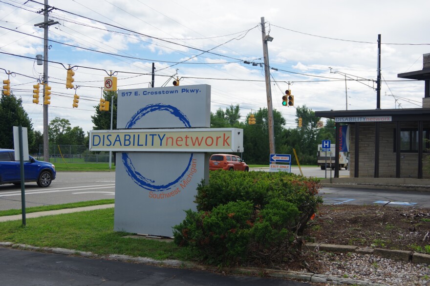 A sign at least five feet high on a green lawn says "Disability Network" with a circle logo. There are bushes and traffic lights nearby. The organization's building is visible behind the sign. It has sandstone walls.
