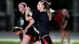A young girl with a blonde ponytail rears back her right hand to throw a football. It's nighttime. She's wearing a dark uniform shirt and shorts and has a belt with red flags tied around her waist. 