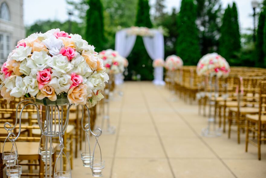 Aisles of wooden chairs and bouquets of flowers lead to an arch draped in sheer cloth. Shrubbery is in the background.