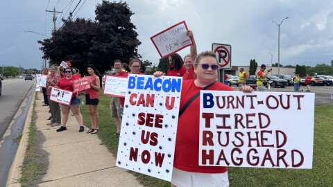 People in red shirts picketing along Broadway Street in Three Rivers. The woman in sun glass featured in the photo holds two red and blue signs that read "Beacon can u see us now" and "Burn-out, Tired, Rushed, Haggard." 