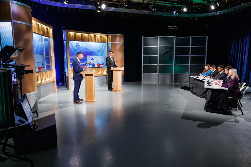 Two men in suits stand at podiums in front of a panel of five people at a TV studio