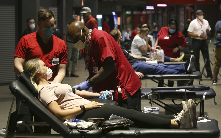 A row of people in masks lay on beds as people in red shirts draw their blood.