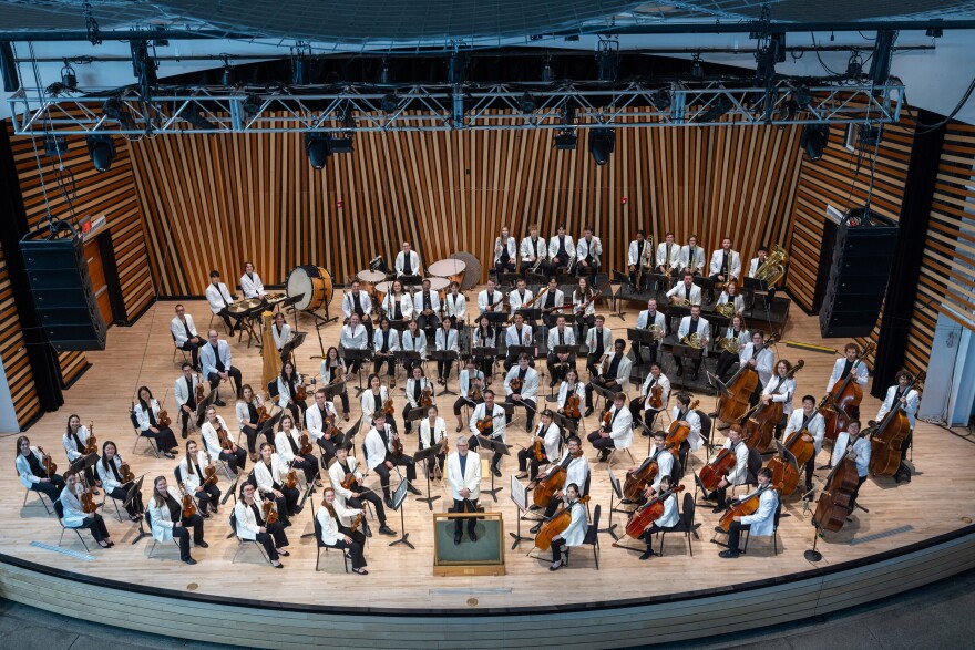 About 80 musicians who are part of the National Repertory Orchestra, are seated on a wooden stage behind the conductor. They all wear black pants and white jackets and are holding their instruments.