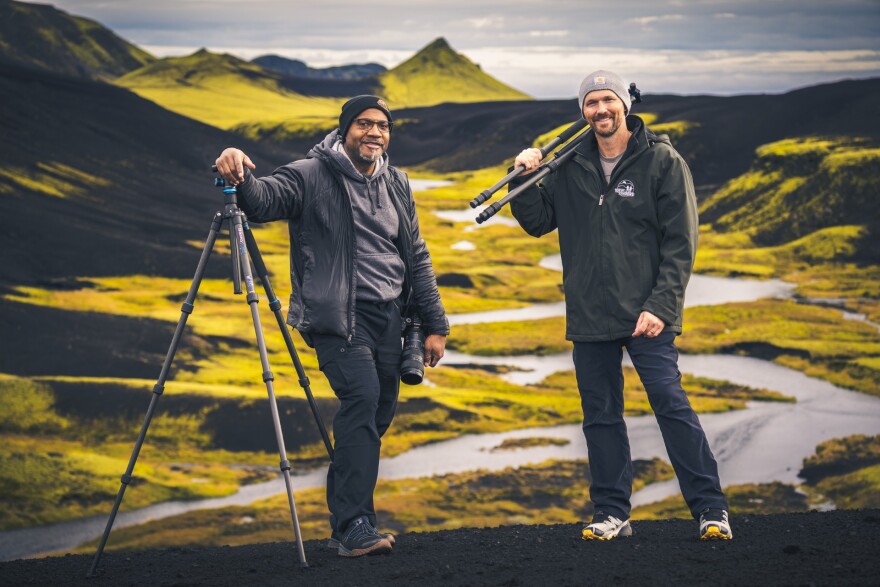 Two men pose with camera gear in a mountain landscape.