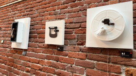 A brick wall with three white boards hanging from it. Each has found objects connected to them as examples of assemblage art.