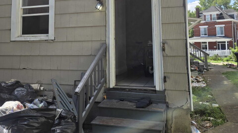 A rear entrance to a single family home. The door and a window are wide open and trash is piled on the ground.