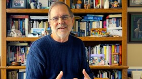 Speaking in front of a bookcase, a man smiles and weaves his fingers together