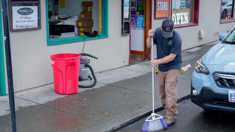 A man sweeps a sidewalk in front of a business missing its store-front window.