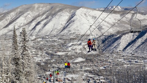Skiers and snowboarders ride up a ski lift with snowy hills and a town in the background.