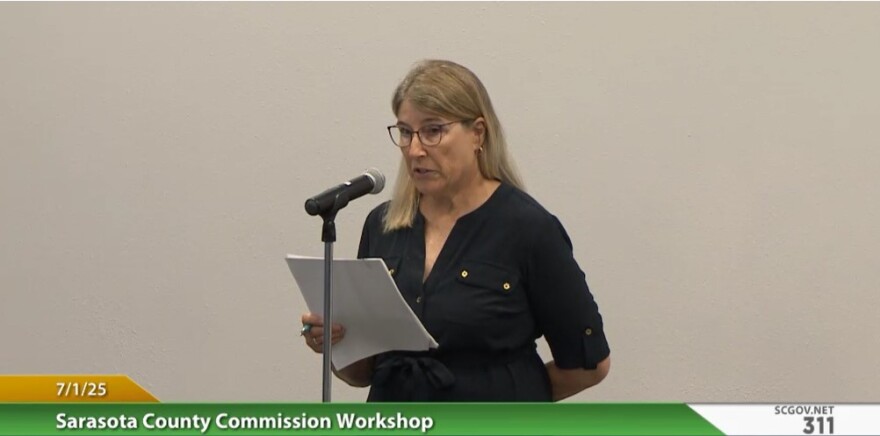 A woman in a black shirt and glasses speaks at a county commission meeting