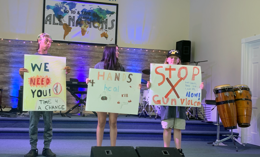 Three campers stand on a stage holding handmade signs with messages against gun violence