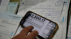 A phone zoomed in on a photo of a high utility bill. The bill is for $2,300.17. Behind the phone are other bills and bookkeeping paperwork. 
