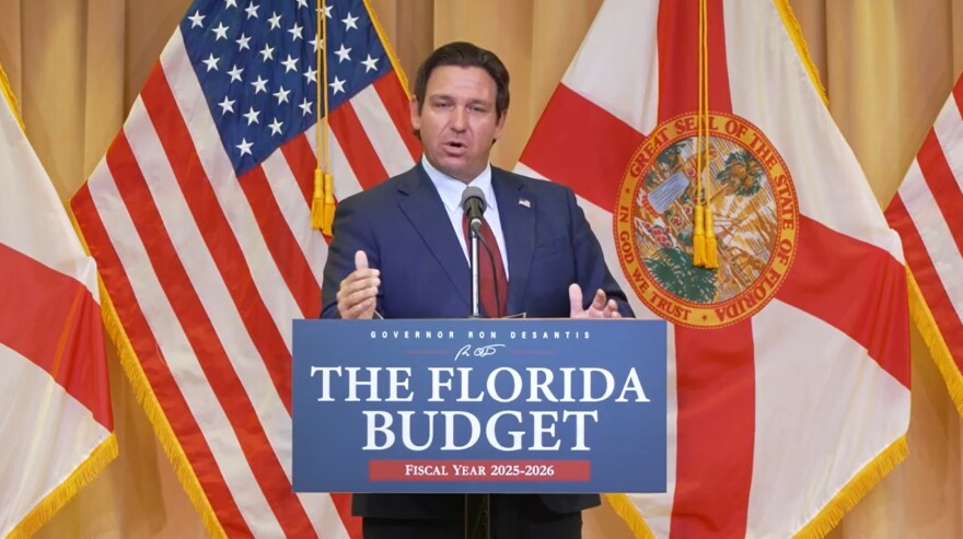 Gov. Ron DeSantis speaks during a press conference about approving a new state budget for the 2025-2026 fiscal year on June 30, 2025. He stands in a blue suit and red tie with the American and Florida flags behind him. 
