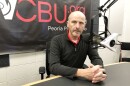 Peoria City Manager Patrick Urich sits at a table in front of a microphone in the WCBU master studio, with the station's brand logo on a banner in the background.  
