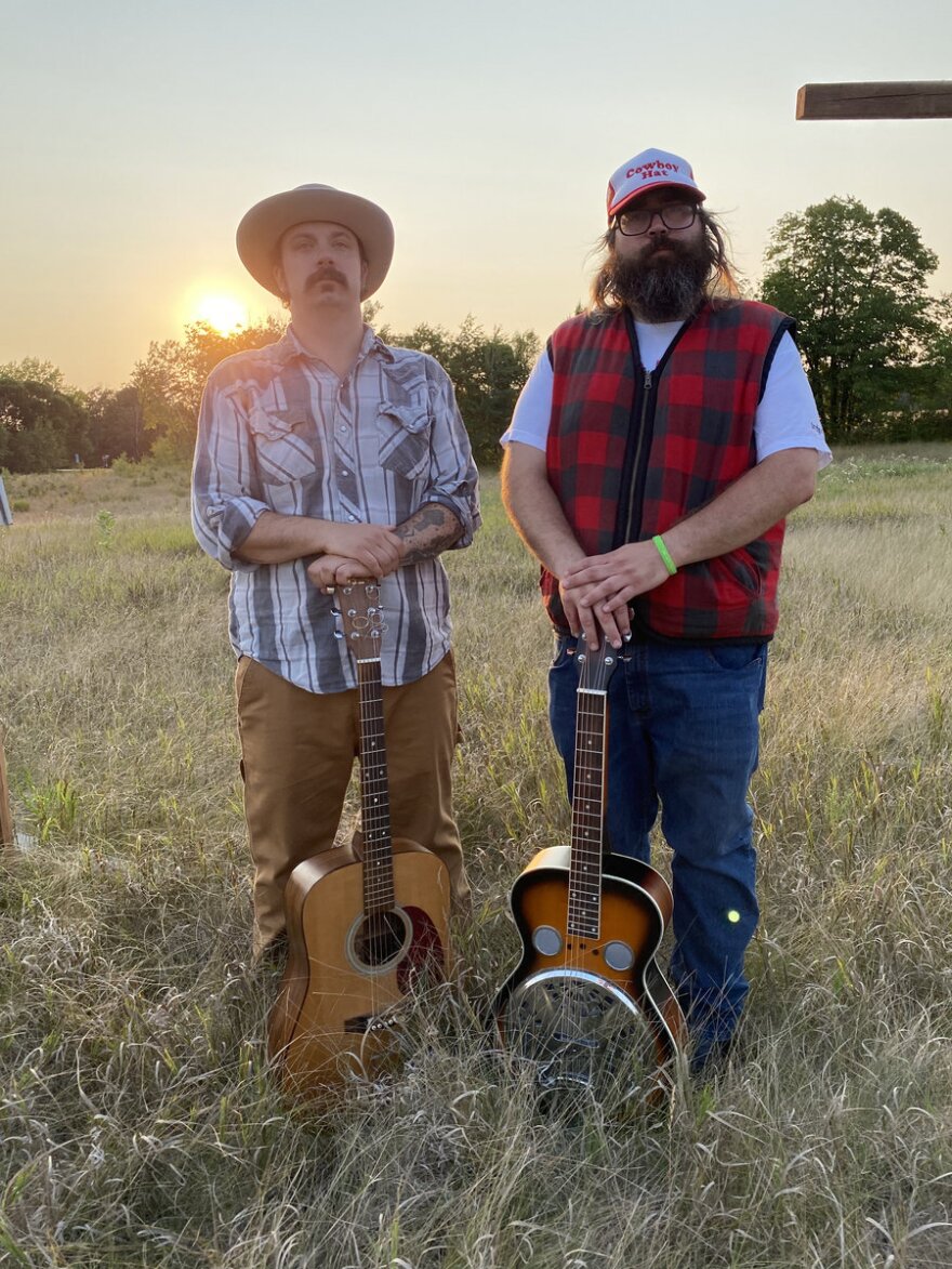 Two men standing in a field with their hands resting on guitars. The sun is setting behind them.