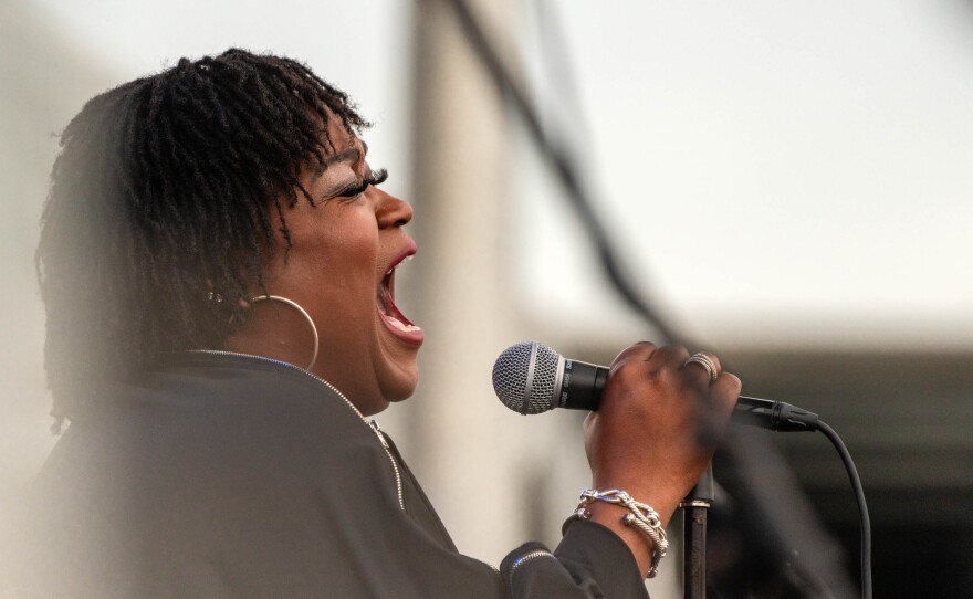 Shemekia Copeland singing into a microphone. She is wearing a black dress and a metallic bracelet.