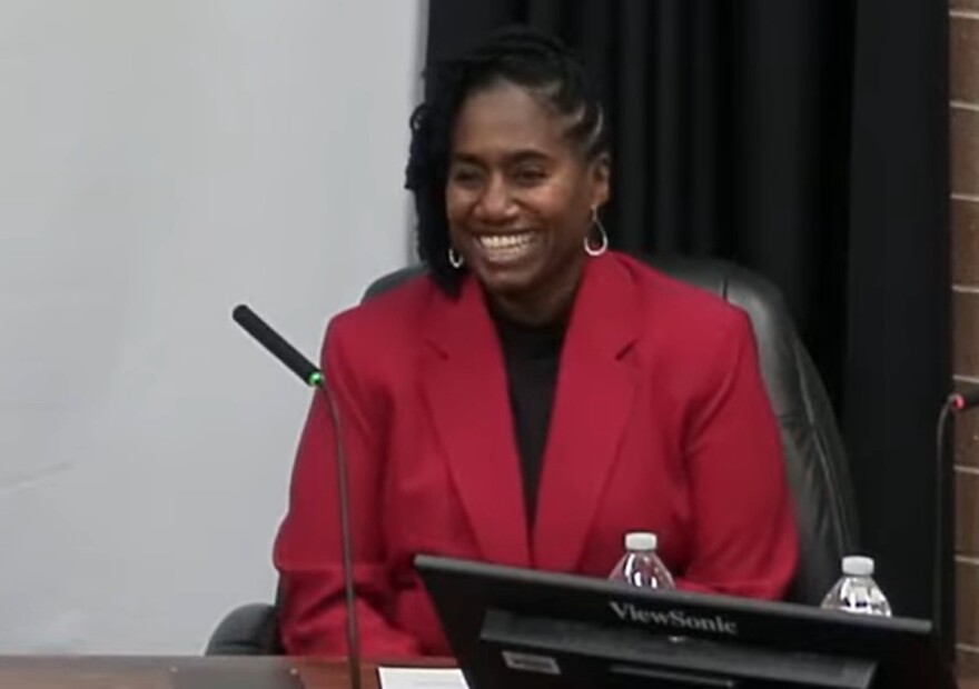 Woman seated at table with microphone and wearing a red jacket and black blouse