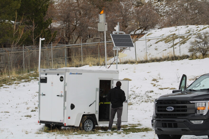 A man stands in front of a small trailer in a snowy field, looking up at a device emitting flames