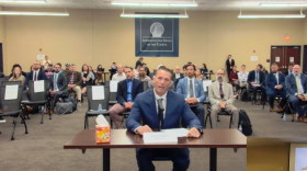 Man sitting at desk speaking into microphone wearing blue suit coat, small crowd of people in audience behind.