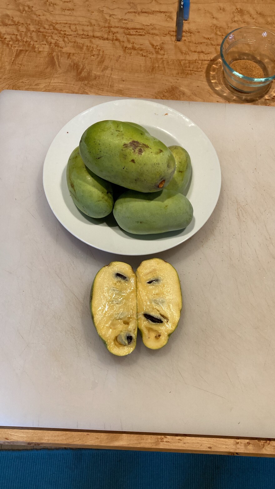 An overhead view of a cutting board with a plate, piled high with green, mango-shaped paw paw fruits. In the foreground, a sliced paw paw fuit reveals its soft, yellow flesh and large, dark magenta seeds.