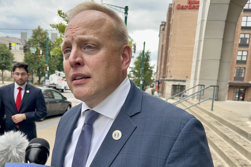 Indiana Solicitor General Thomas Fisher stands outside of the Monroe County Court building after a hearing, speaking to reporters. Fisher is a White man with light blonde hair. He's wearing a blue suit, a white shirt and a blue tie.