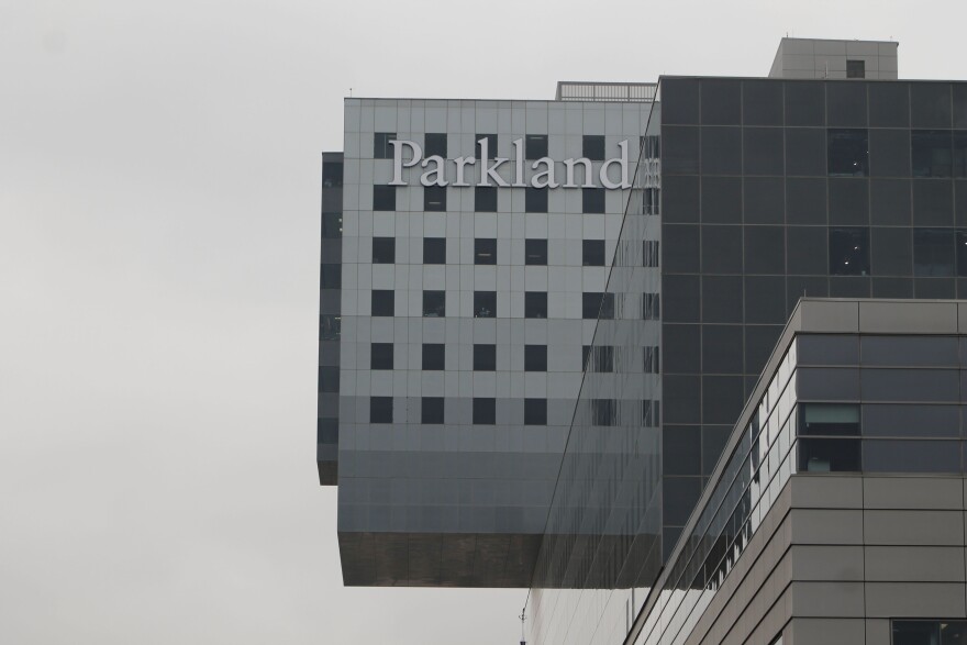 Parkland Hospital on a very overcast day. One top of a dark glass structure is a gray structure with a sign that reads "Parkland" in the upper left corner. 