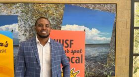Black man with a beard and blue sportcoat smiling into the camera with a peach WUSF banner behind him