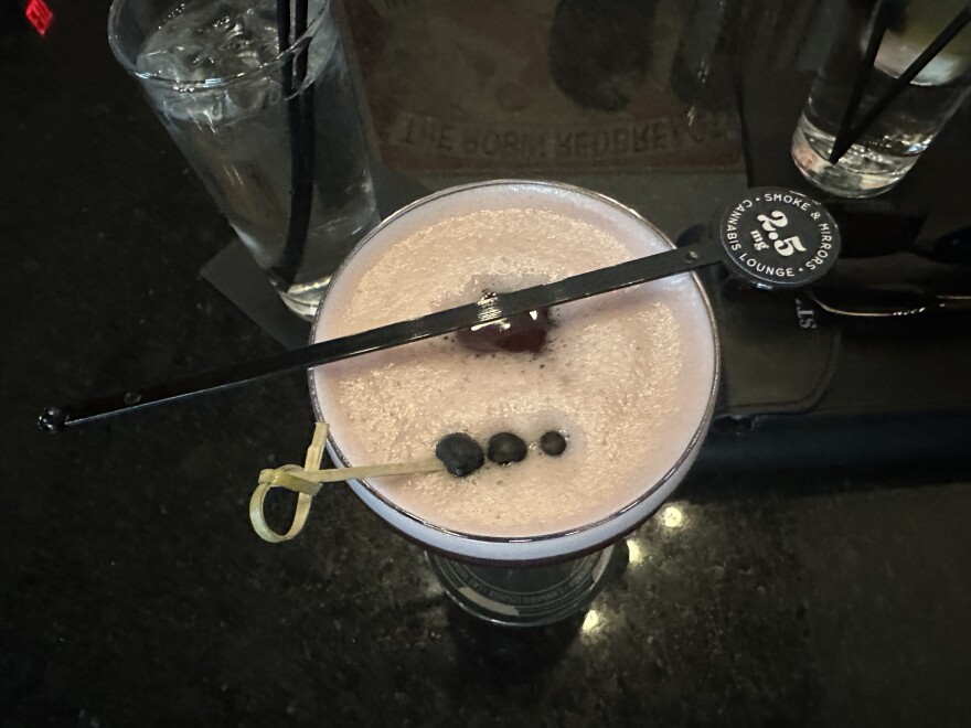 A frothy cocktail viewed from above with a skewer with three berries. Across the glass is a stirrer with a circular top that reads "2.5 mg, Smoke and Mirrors Cannabis Lounge".