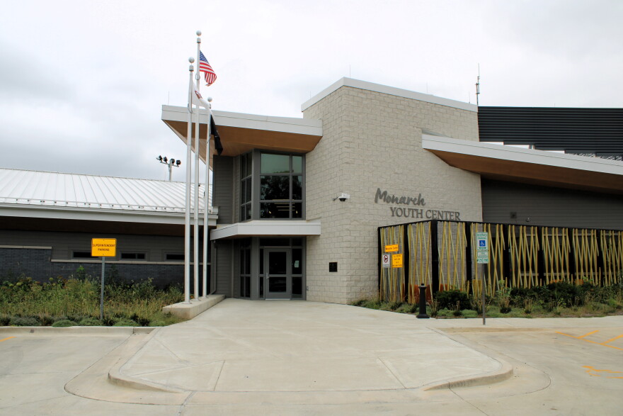 The exterior entrance of the Monarch Youth Center, featuring a modern building with a light stone facade, a concrete walkway, and an American flag flying on a pole.