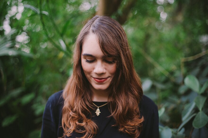 Woman with long brown hair smiling and looking down with trees behind her