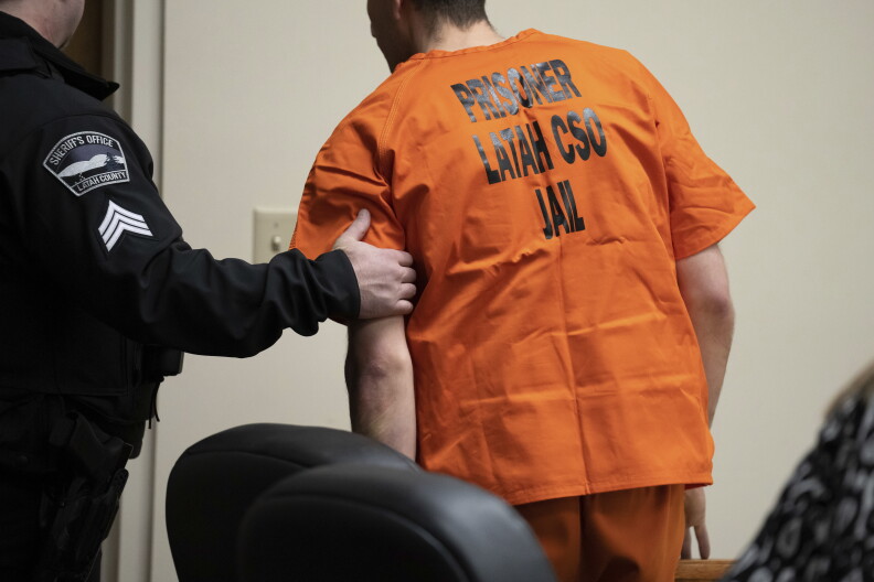 A man wearing an orange jumpsuit with the words "Prisoner Latah CSO Jail" written in black letters in the back is not facing the camera. His face is not visible. A police officer holds his left arm as he is being escorted.