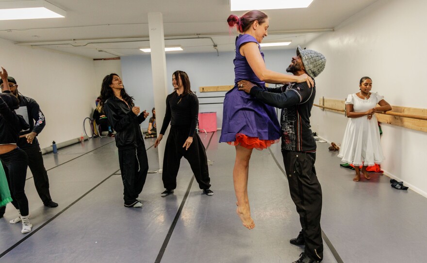 A man lifts a dancer wearing a purple and red dress up into the air