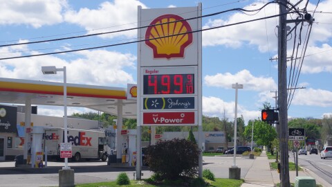 A Shell gas station sign next to a busy road displays the price for gas; $4.99