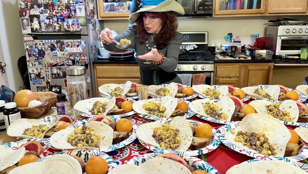 A table is covered with paper plates filled with food. An older woman with long brown hair and a large hat spoons food onto a plate. 