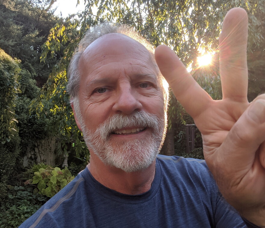 Older man with gray hair and beard smiles outdoors, holding up a peace sign with his fingers. Sunlight shines through trees in the background.