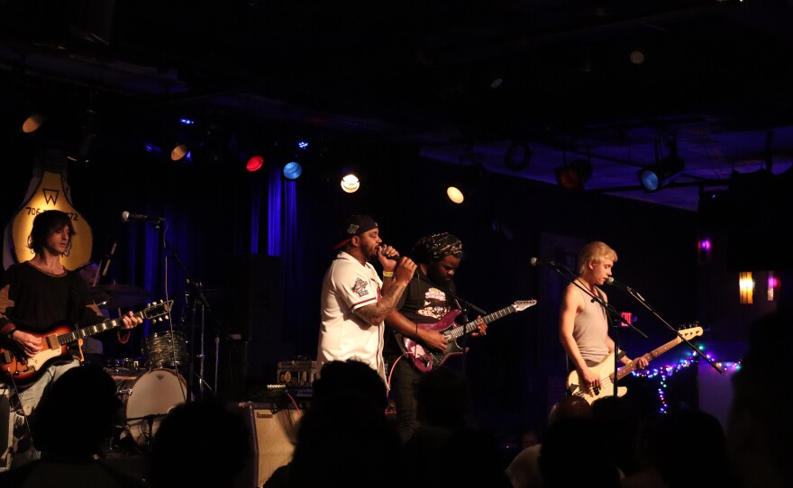 Four musicians perform on a dark indoor stage.